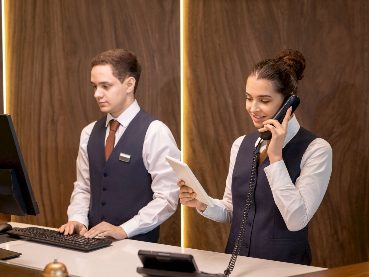 Two hotel receptionists work at a desk; one types on a computer while the other answers a phone and holds a clipboard.