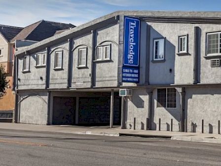 The image shows a Travelodge hotel building on a clear day, with a blue sign displaying the hotel's name and some accommodations listed.