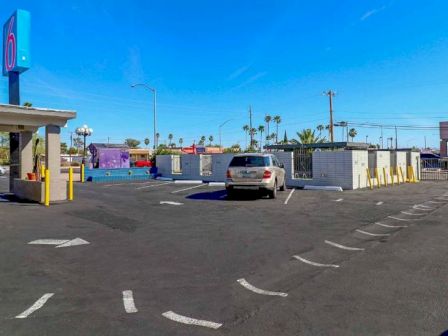 The image shows a parking lot with a few structures, a car, and directional arrows. The sky is clear and blue. There are some palm trees in the distance.