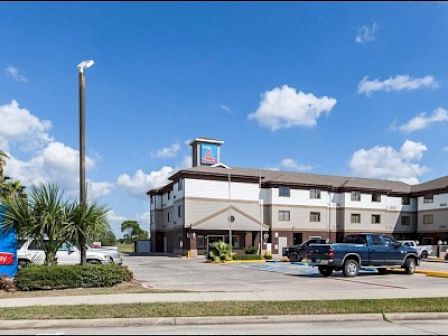 The image shows a Studio 6 Extended Stay hotel with a parking lot, some vehicles, and a clear blue sky with scattered clouds.