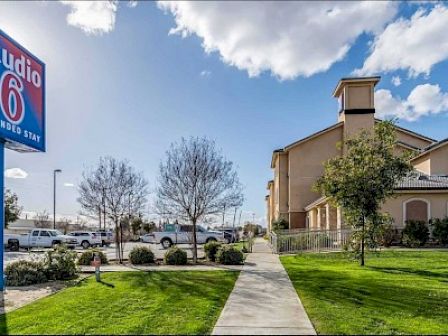 The image shows a Studio 6 Extended Stay hotel building, a blue sign, a pathway, green lawns, and parked vehicles under blue skies with fluffy clouds.