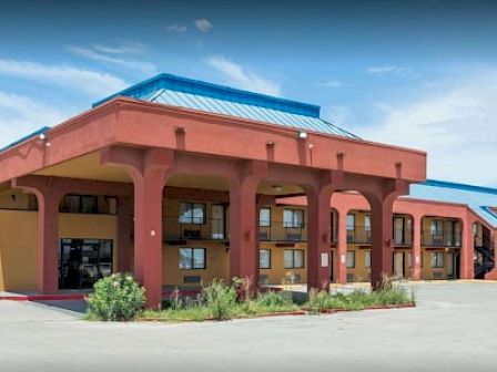 The image shows a two-story building, likely a hotel or motel, with blue roofing and a large parking area in front.