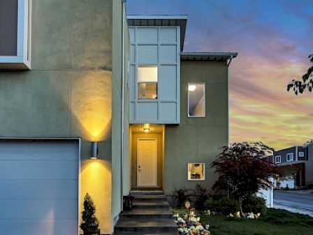 A modern house with illuminated entrance, large windows, and a garage, against a colorful sunset sky. A small garden decorates the front yard.