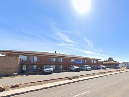 The image shows a two-story motel with a few vehicles parked outside under a clear sky. There is a sign to the right of the motel.