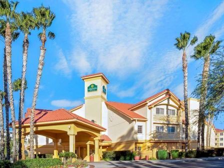 This image shows a hotel building with cream-colored walls and red roof tiles, surrounded by palm trees and a clear blue sky in the background.