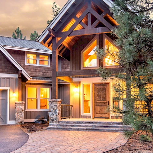 A beautiful, rustic two-story house with large windows and warm lights, nestled among tall trees, featuring a stone pathway and a two-car garage.