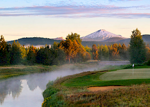 A serene landscape featuring a golf course, a river, dense trees, and a snow-capped mountain in the background under a colorful sky.