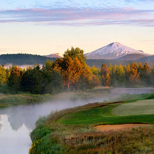 A serene landscape featuring a golf course, a river, dense trees, and a snow-capped mountain in the background under a colorful sky.