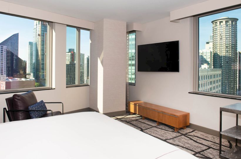 A modern hotel room with large windows, a bed, chair, TV, wooden cabinet, and city skyline view.
