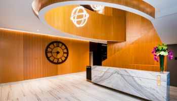 A modern lobby with a marble reception desk, wooden spiral staircase, large clock on the wall, and a vase with flowers on the desk.
