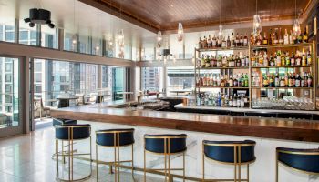 A modern bar with a wooden counter, stylish seating, and a shelf full of various liquor bottles, situated in a room with large windows and city views.