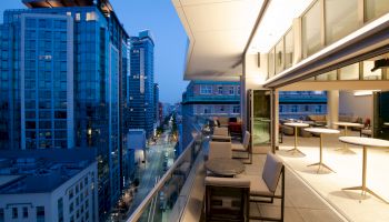 A modern cityscape at dusk, featuring high-rise buildings and a sleek balcony with seating and tables, overlooking a lit-up street.