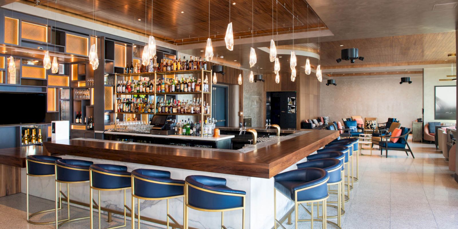 This image shows a modern bar with a wooden counter, blue chairs, a well-stocked bar shelf, and stylish lighting fixtures, with seating areas around.