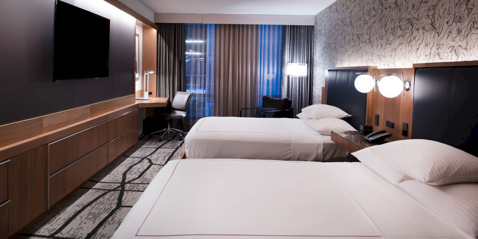 This image shows a modern hotel room with two double beds, a flat-screen TV, a work desk with a chair, and contemporary decor.