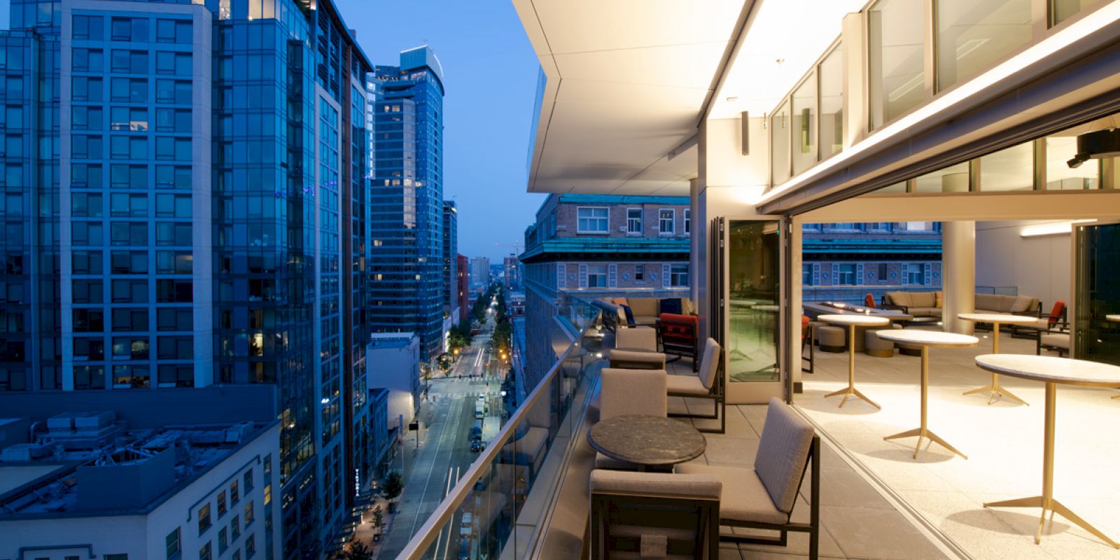 This image shows a modern urban balcony with tables and chairs, overlooking city buildings during evening time.