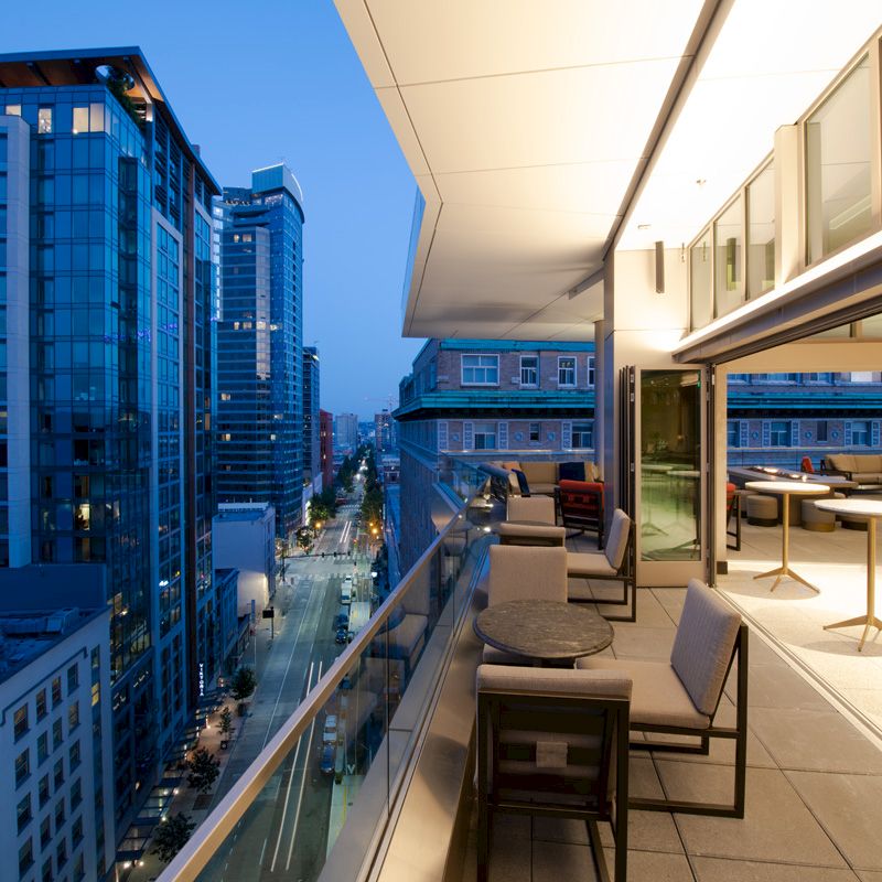 A modern cityscape at dusk is seen from a high-rise building's balcony with lounge chairs and tables overlooking illuminated buildings and streets.