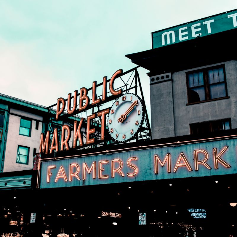 The image shows a public market with a large clock and neon signs reading "Public Market" and "Farmers Market" on a building façade.