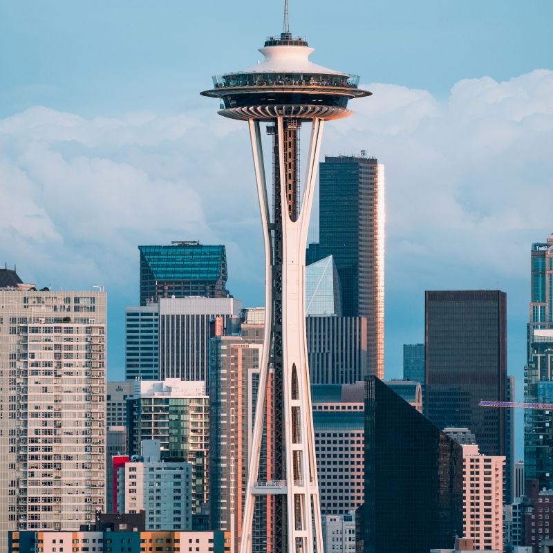 The image shows the Seattle skyline with the iconic Space Needle in the foreground, surrounded by various high-rise buildings under a partly cloudy sky.