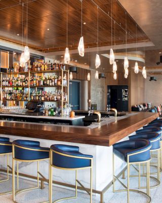 The image shows a modern bar with blue barstools, a large selection of drinks on shelves, hanging lights, and a cozy seating area in the background.