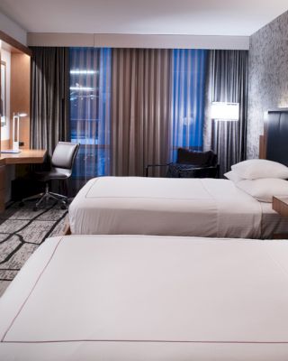 A modern hotel room with two beds, a wall-mounted TV, a desk with a chair, and stylish decor, lit by lamps and natural light from the window.