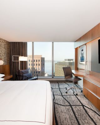 A modern hotel room with a large bed, desk, and flat-screen TV. The window offers a view of nearby buildings and possibly water in the distance.