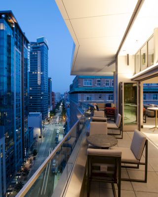 A modern cityscape view from a balcony with sleek furniture, looking out over tall buildings and a street below at dusk.