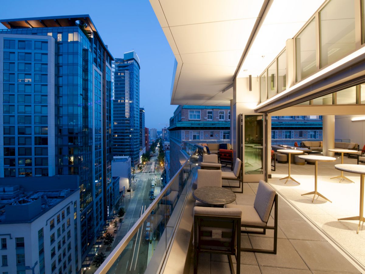 A modern balcony with seating, city buildings, and a street view at dusk. The indoor space is visible through glass, featuring tables and chairs.