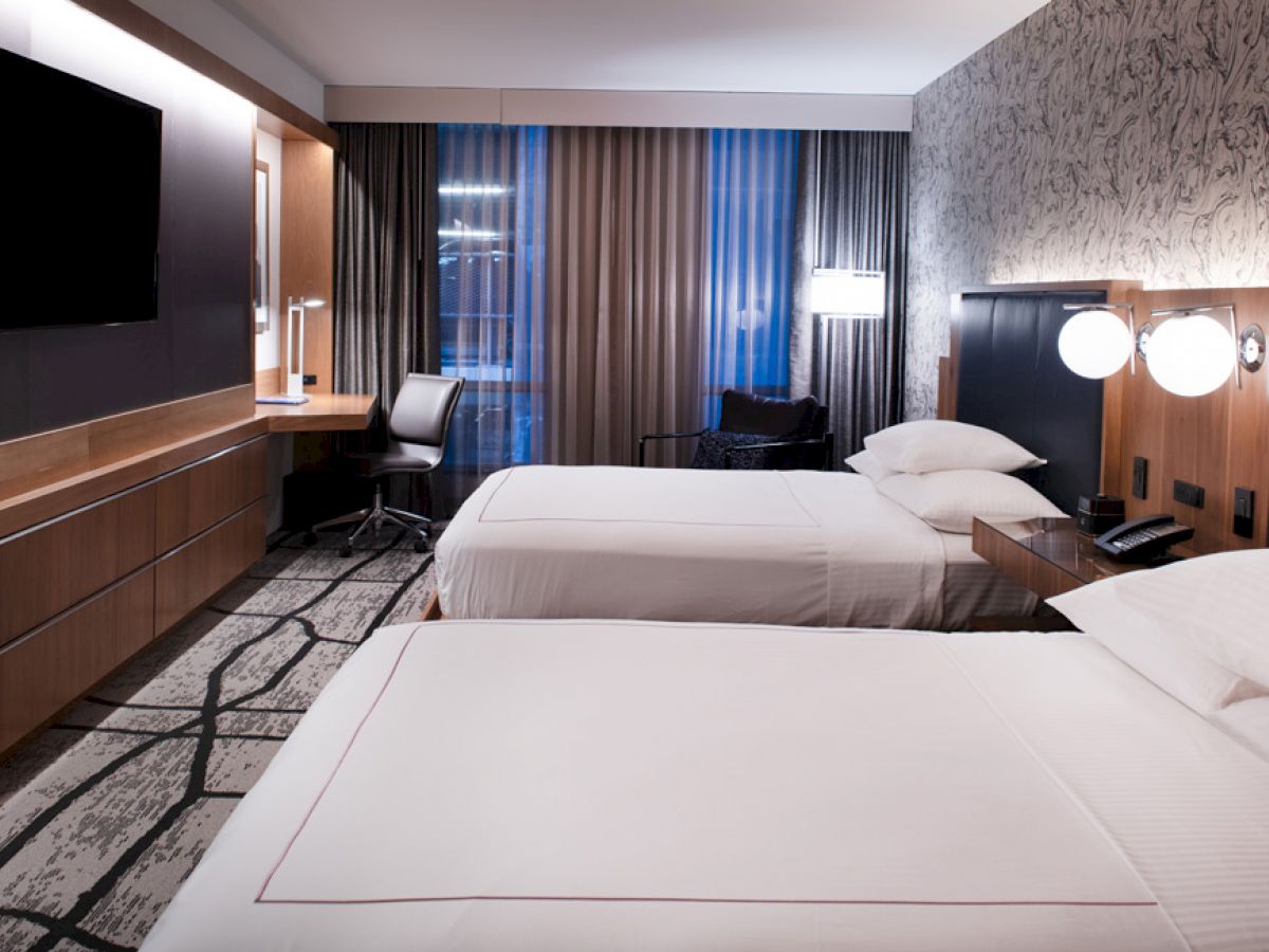 A modern hotel room with two neatly made double beds, a flat-screen TV, a desk with a chair, and large windows covered by curtains.