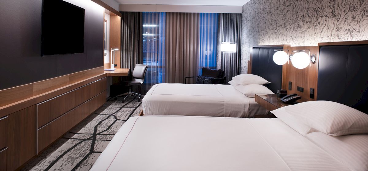 The image shows a modern hotel room with two neatly made beds, a flat-screen TV, a desk with a chair, and contemporary lighting.
