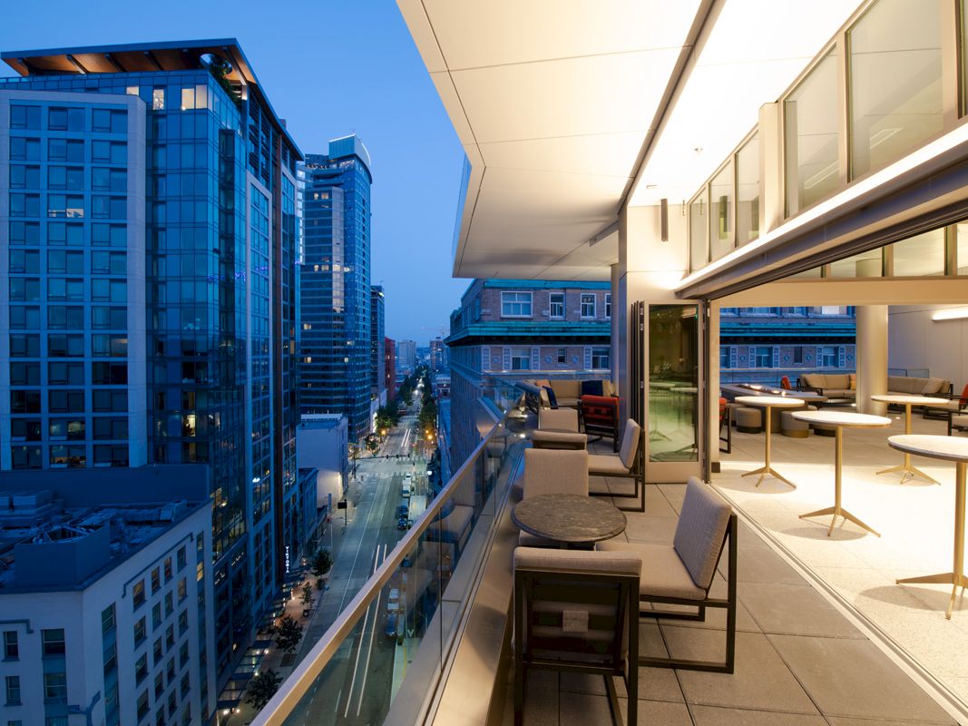 A modern balcony view at dusk featuring cityscape high-rise buildings, stylish furniture, glass railings, and lighting from the interior space.
