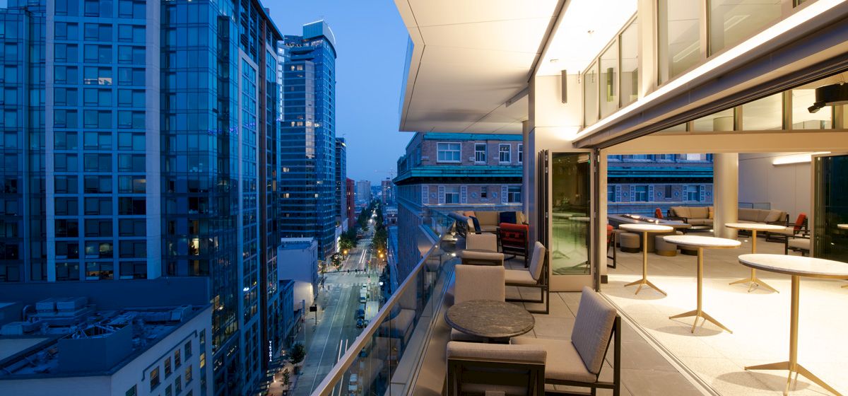 A modern balcony view at dusk featuring cityscape high-rise buildings, stylish furniture, glass railings, and lighting from the interior space.