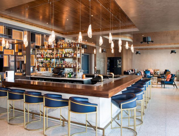 This image shows a modern bar with a well-stocked liquor shelf, blue cushioned bar stools, pendant lights, and seating areas in the background.