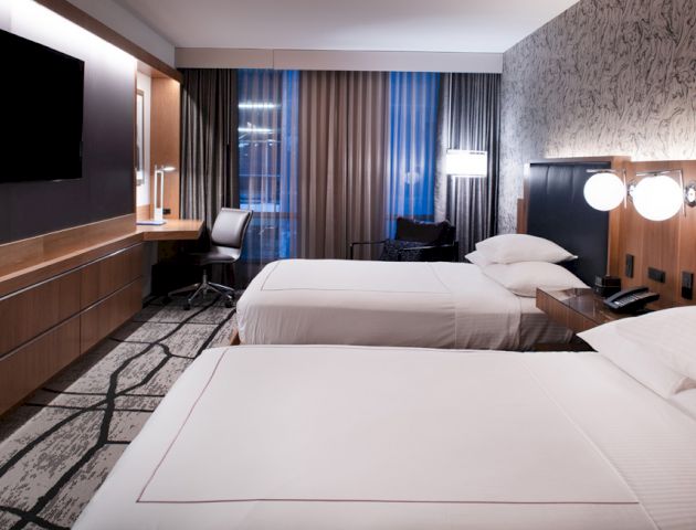 A modern hotel room with two beds, a wall-mounted TV, a desk with a chair, lamps, and curtains with natural light filtering through the window.