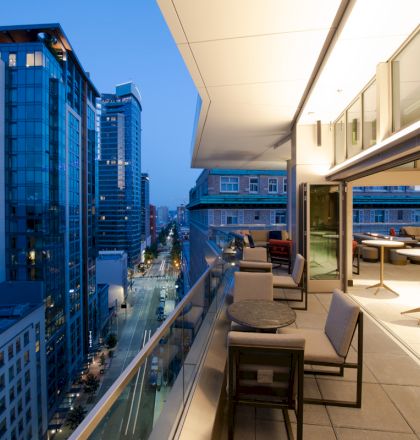 Outdoor patio with modern furniture overlooking a city street and high-rise buildings at dusk, featuring tables, chairs, and warm lighting.