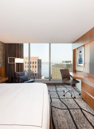 A modern hotel room features a king-size bed, large wall-mounted TV, desk, and chair, with a city view through floor-to-ceiling windows.