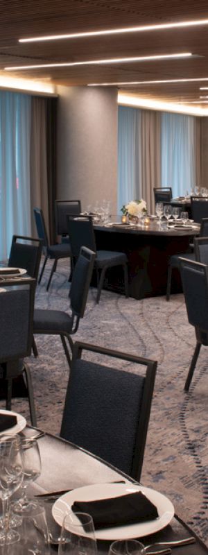A formal dining setup in an elegant room with round tables, black chairs, tableware, and flower centerpieces, illuminated by ceiling lights.