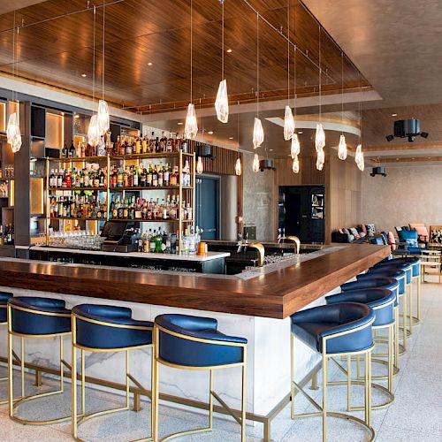 A modern bar features a large counter with blue and gold stools, shelves stocked with bottles, and hanging lights, creating an inviting atmosphere.