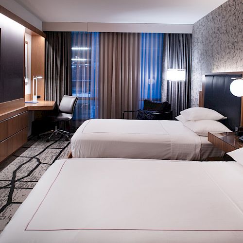 The image shows a modern hotel room with two neatly made beds, a desk by the window, a flat-screen TV, and elegant lighting on the wall.