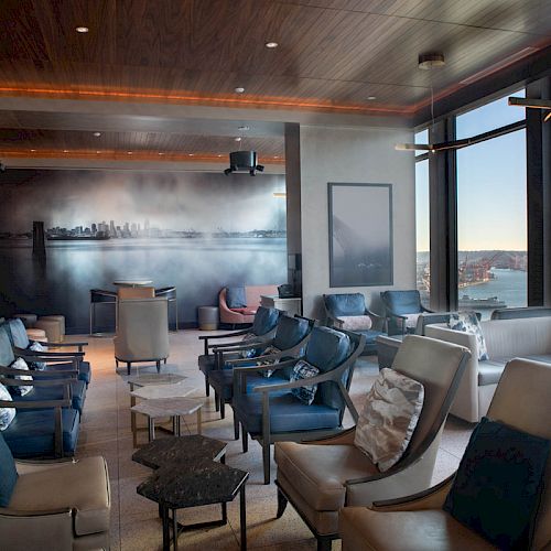 The image shows a lounge area with multiple chairs facing large windows with an ocean view and a Ferris wheel in the distance.
