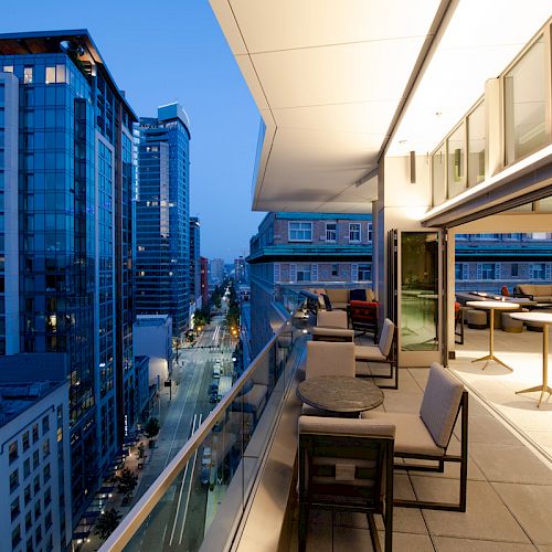 A cityscape at twilight featuring modern high-rise buildings and a balcony with seating overlooking a street.