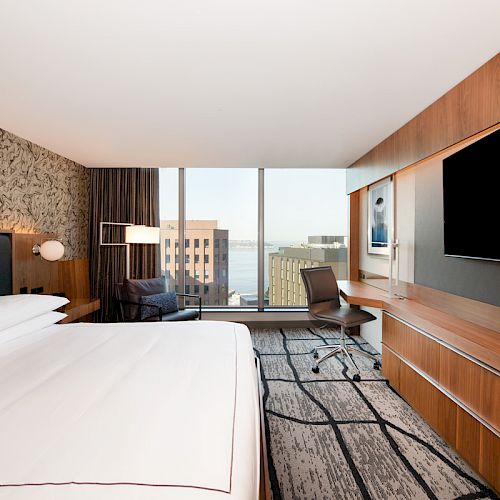 The image shows a modern hotel room with a large bed, wall-mounted TV, desk, chair, and windows with a cityscape view.