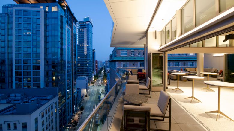 An open balcony view from a modern building with seating areas, overlooking a city street lined with tall buildings at dusk.