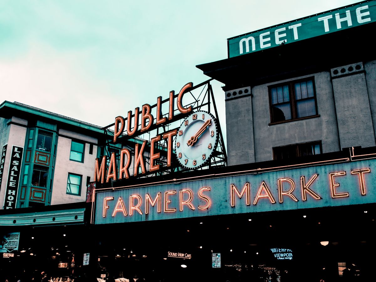 The image shows a public farmers market with neon signs and a clock on the facade of a building. The market atmosphere is lively and inviting.
