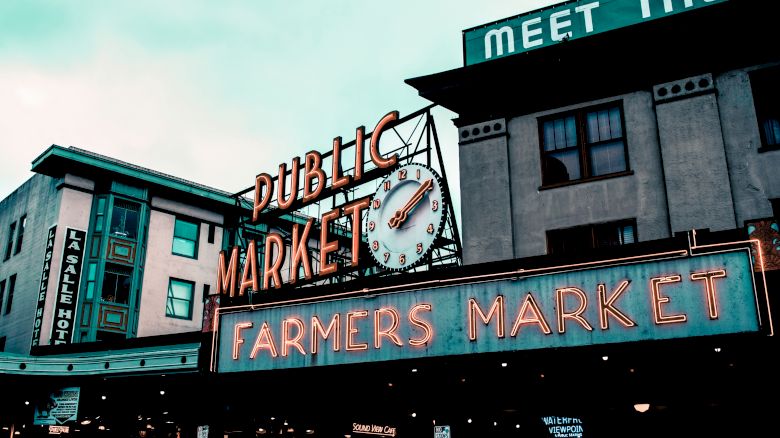 The image shows a public farmers market with neon signs and a clock on the facade of a building. The market atmosphere is lively and inviting.