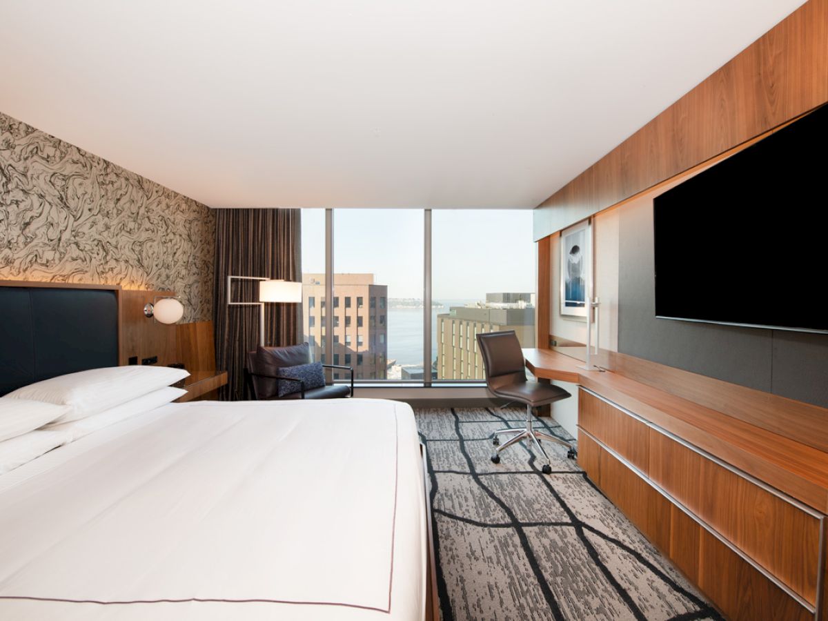 A modern hotel room with a large bed, desk, wall-mounted TV, and a view of buildings and water through a wide window, showcasing contemporary decor.