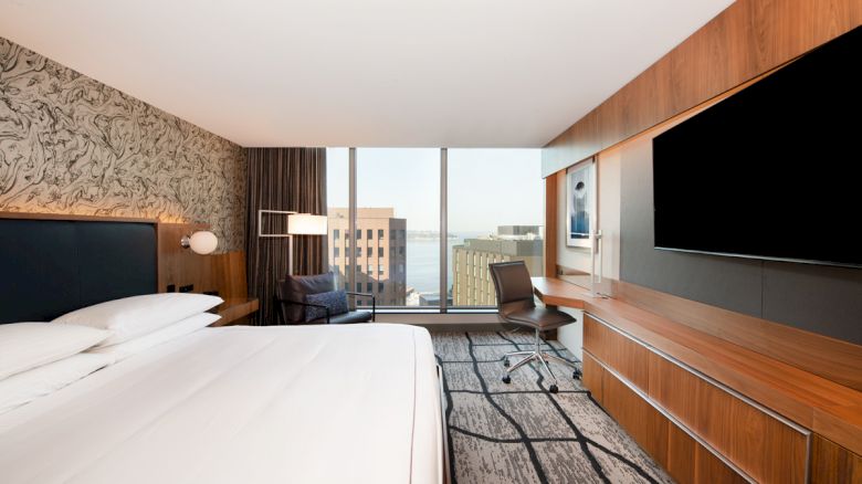 A modern hotel room with a large bed, desk, wall-mounted TV, and a view of buildings and water through a wide window, showcasing contemporary decor.