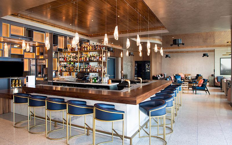 The image shows a modern bar with a central counter, blue barstools with gold accents, hanging light fixtures, and a well-stocked shelf of liquor bottles.