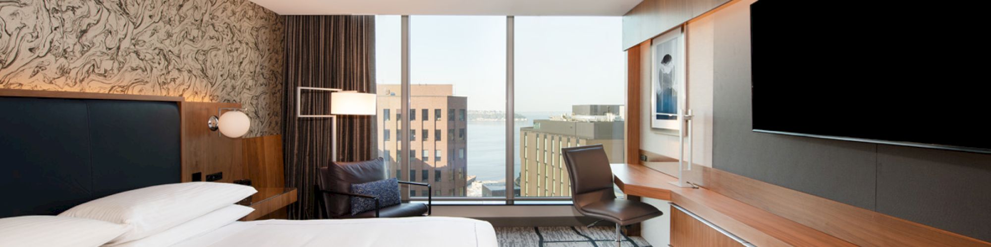 A modern hotel room with a large bed, a wall-mounted TV, a work desk, a chair, and a window offering a city view with buildings.