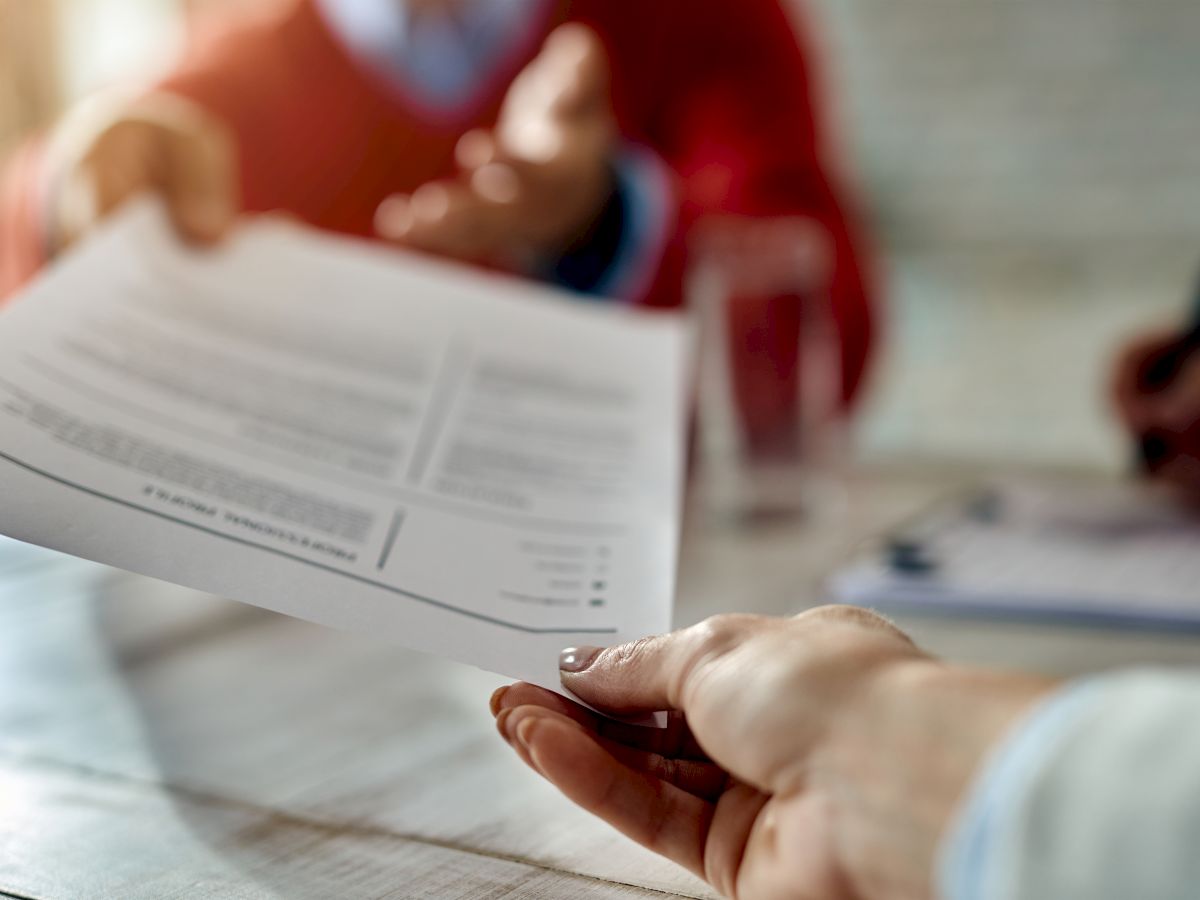 A person is handing over a document to another individual in a professional setting, while a third person is seen writing in the background.